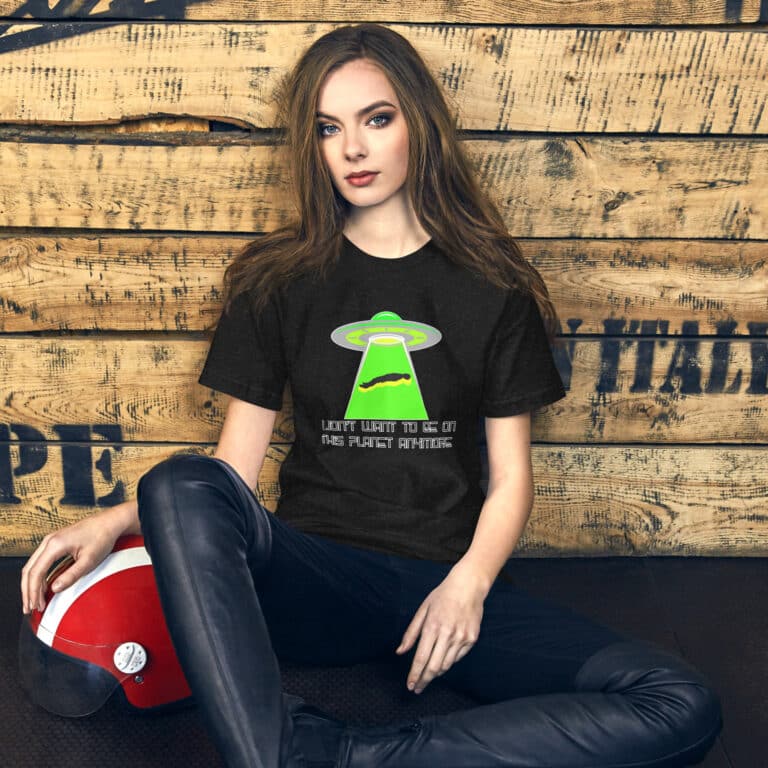 A person sits against a wooden wall, wearing a T-shirt with the product name "I Don't Want To Live On This Planet Anymore," which showcases a green UFO graphic. Beside them on the floor is a red helmet.