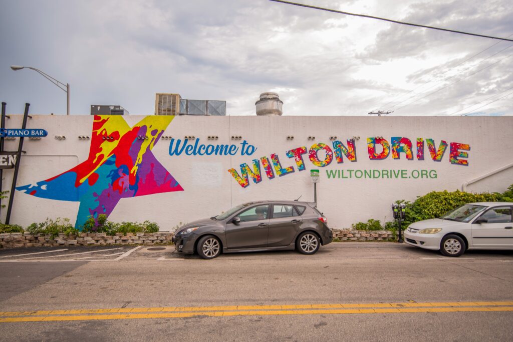 A mural on a white wall reads "Welcome to Wilton Drive" in colorful letters with a rainbow star, highlighting the vibrant spirit of Wilton Drive; two cars are parked in front.