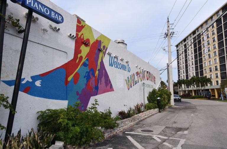 A colorful mural on a white wall reads “Welcome to Wilton Drive,” capturing the vibrant spirit of Wilton Drive beside a street lined with a piano bar sign and an apartment building in the background.