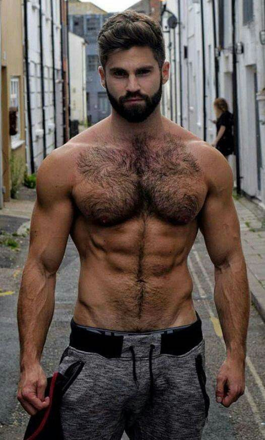 Shirtless man with a muscular build and dark body hair stands in an alleyway wearing grey athletic pants and holding a garment in one hand.