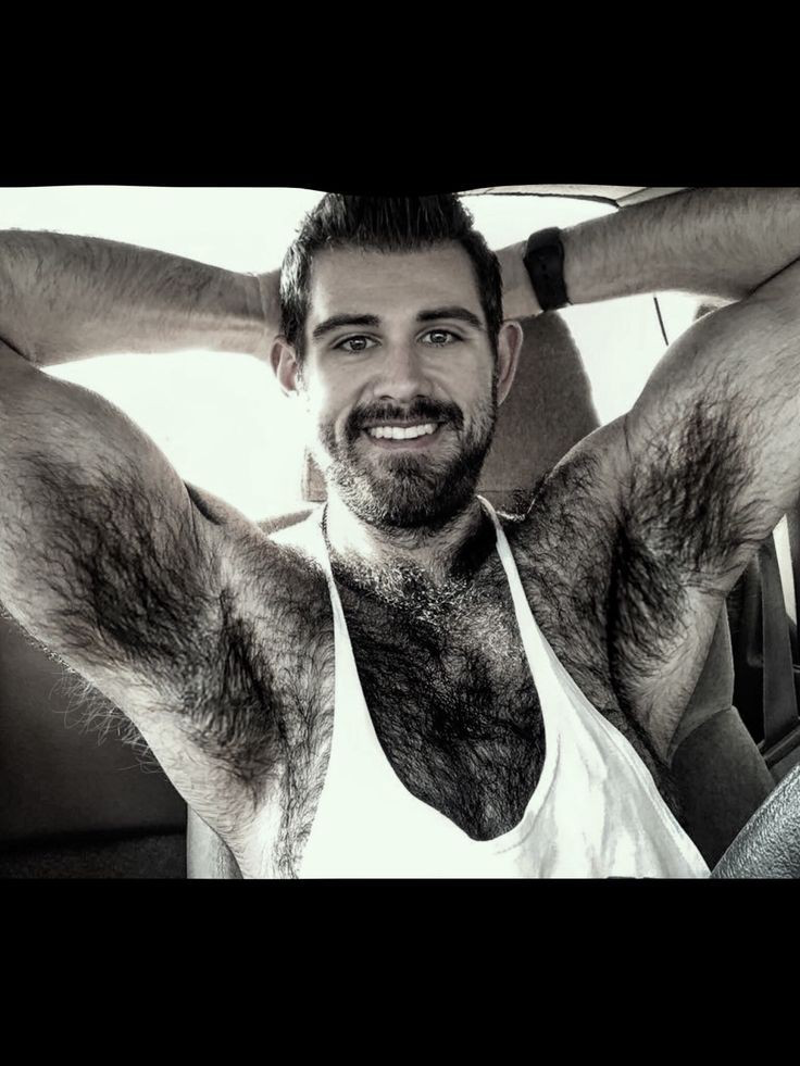 A man with a beard and body hair sits in a car, wearing a sleeveless white tank top and smiling with his arms raised behind his head.