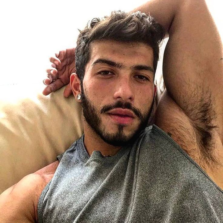 A man with dark hair, body hair, and a beard reclines on a couch with one arm behind his head, wearing a sleeveless gray shirt and an earring.