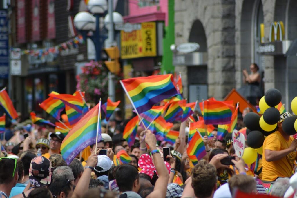 A large crowd waves rainbow flags at a street event in a vibrant city—some hold black and yellow balloons and colorful signs. It’s the perfect spot to visit while exploring the top 10 LGBTQ+ neighborhoods during your travels.