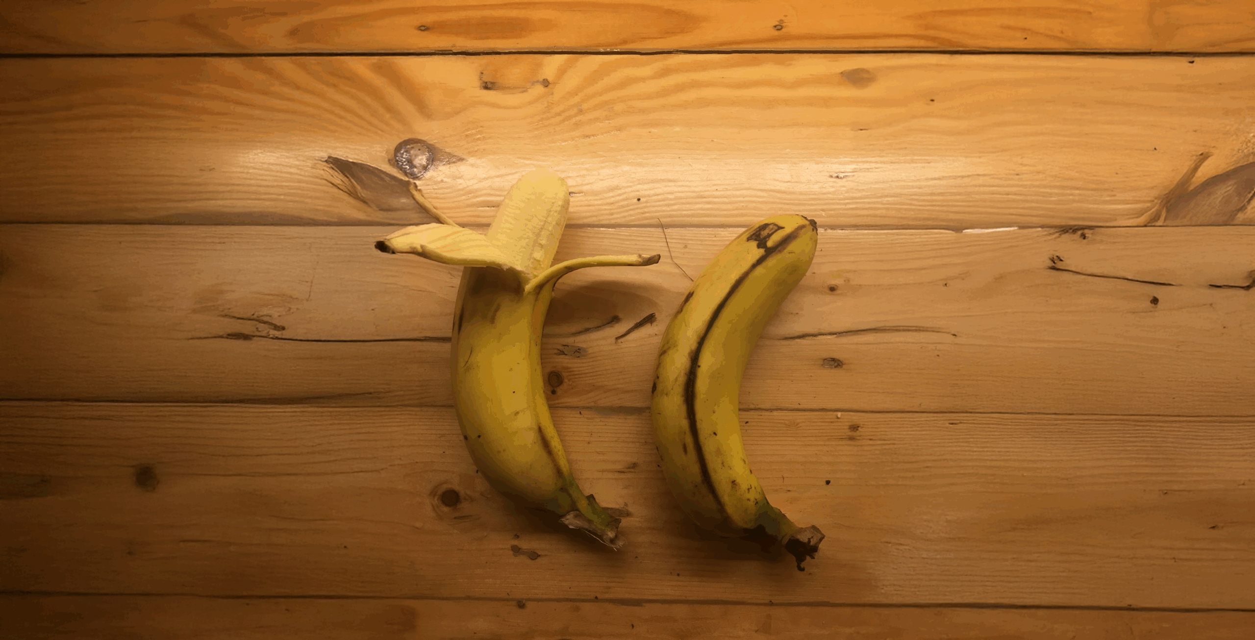 Two bananas on a wooden surface; one is partially peeled open, while the other remains unpeeled.