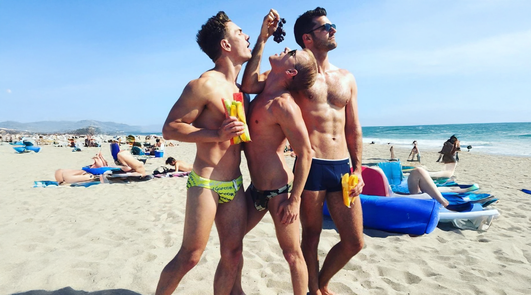 Three men in swimwear stand on a sandy beach, posing playfully with popsicles while other beachgoers relax in the background.