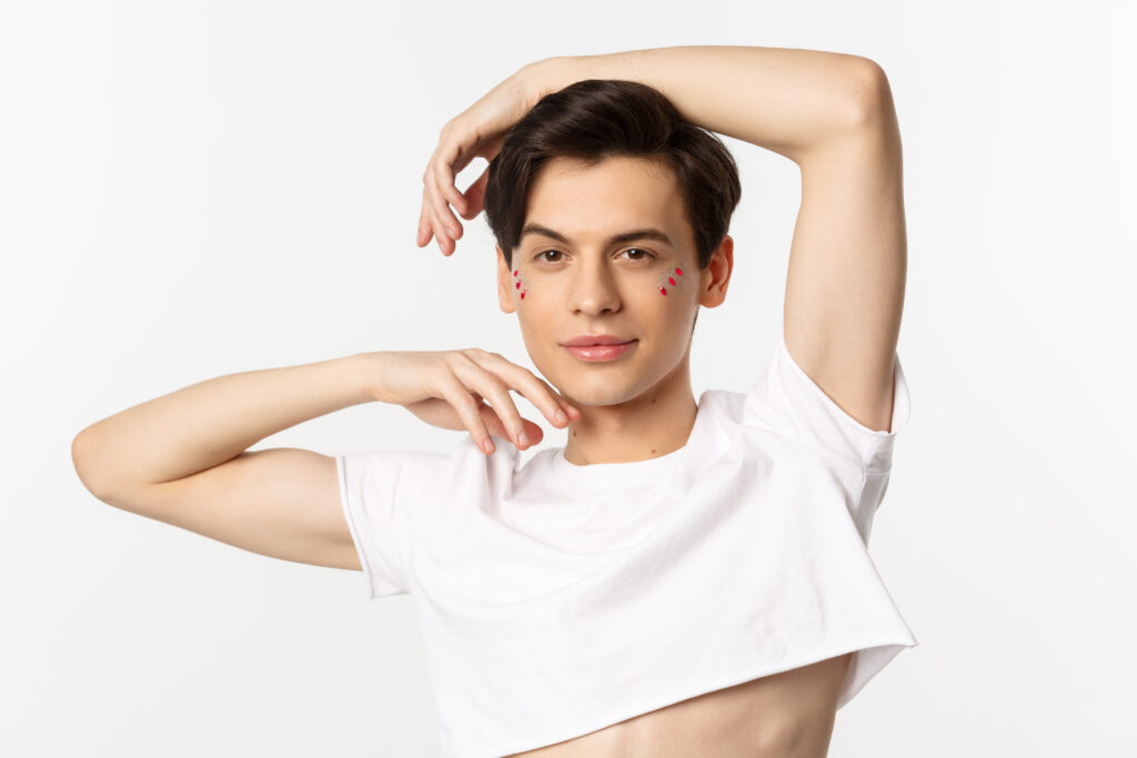Young person in a white crop top poses with one arm raised, small decorative heart stickers on their cheeks, channeling playful vibes that would pair perfectly with the Funniest Gay Shirts collection, all against a plain white background.
