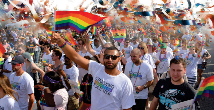 A group of people in white shirts reading Standing Together march in a Florida Pride parade, waving rainbow flags amid colorful confetti.