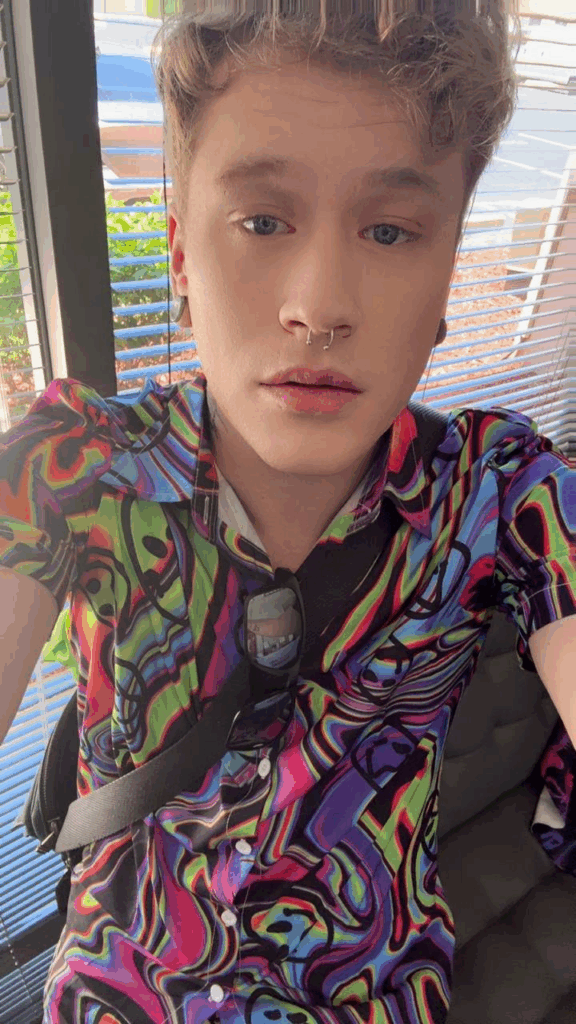 A person with short curly hair and a septum piercing wears a colorful abstract-patterned shirt indoors near a window, getting ready for Stonewall Pride in Wilton Manors, Florida.