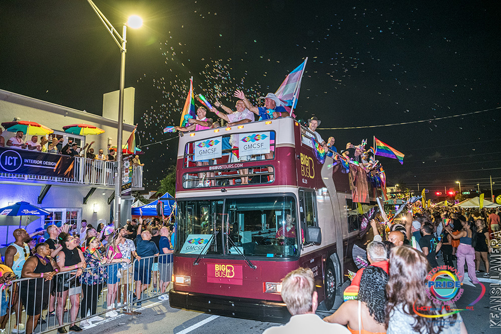 A double-decker bus full of people waving Pride flags and blowing bubbles rolls through a crowded street at night during the Stonewall Pride Glow parade in Florida, with spectators cheering from the sidewalks.